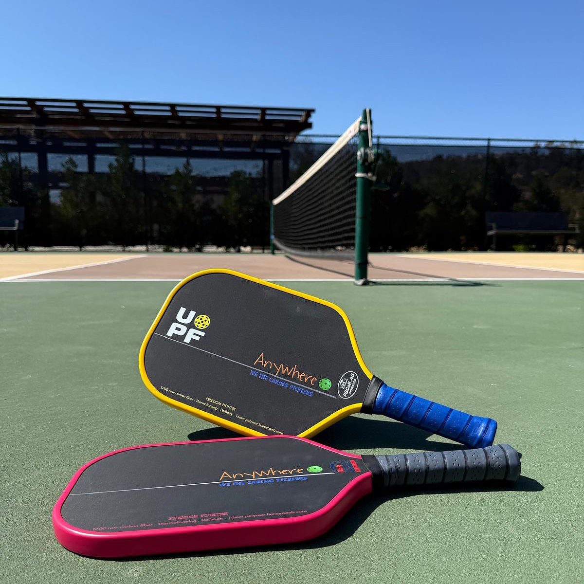 Two Anywhere Freedom Fighter pickleball paddles, one yellow and one red, rest on an outdoor court near the net.