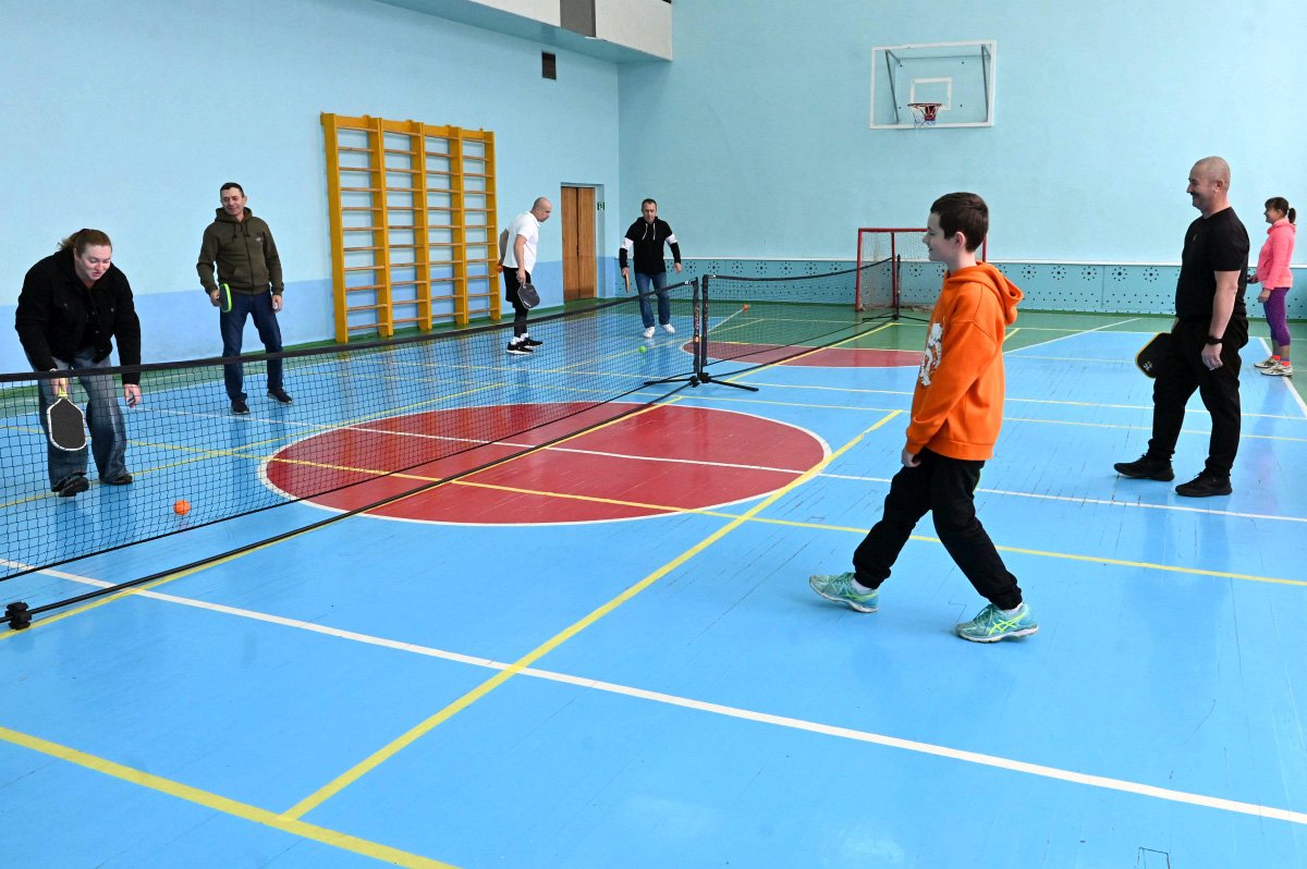 Ukrainian men, women, and children play pickleball together in an indoor gym.