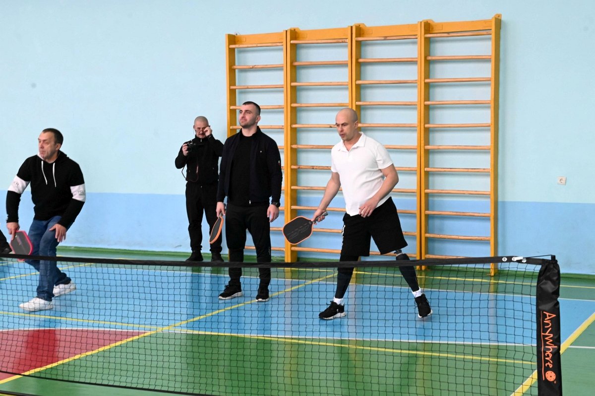 Ukrainian man with a prosthetic leg plays pickleball indoors with other participants.